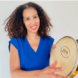 Melissa Mister is standing in front of a white background wearing a blue blouse. She is holding a tambourine and smiling at the camera.